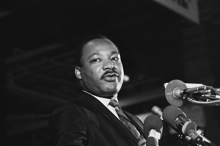 Black and white photo of Martin Luther King Jr. looking off to the side as several mics sit in front of him.