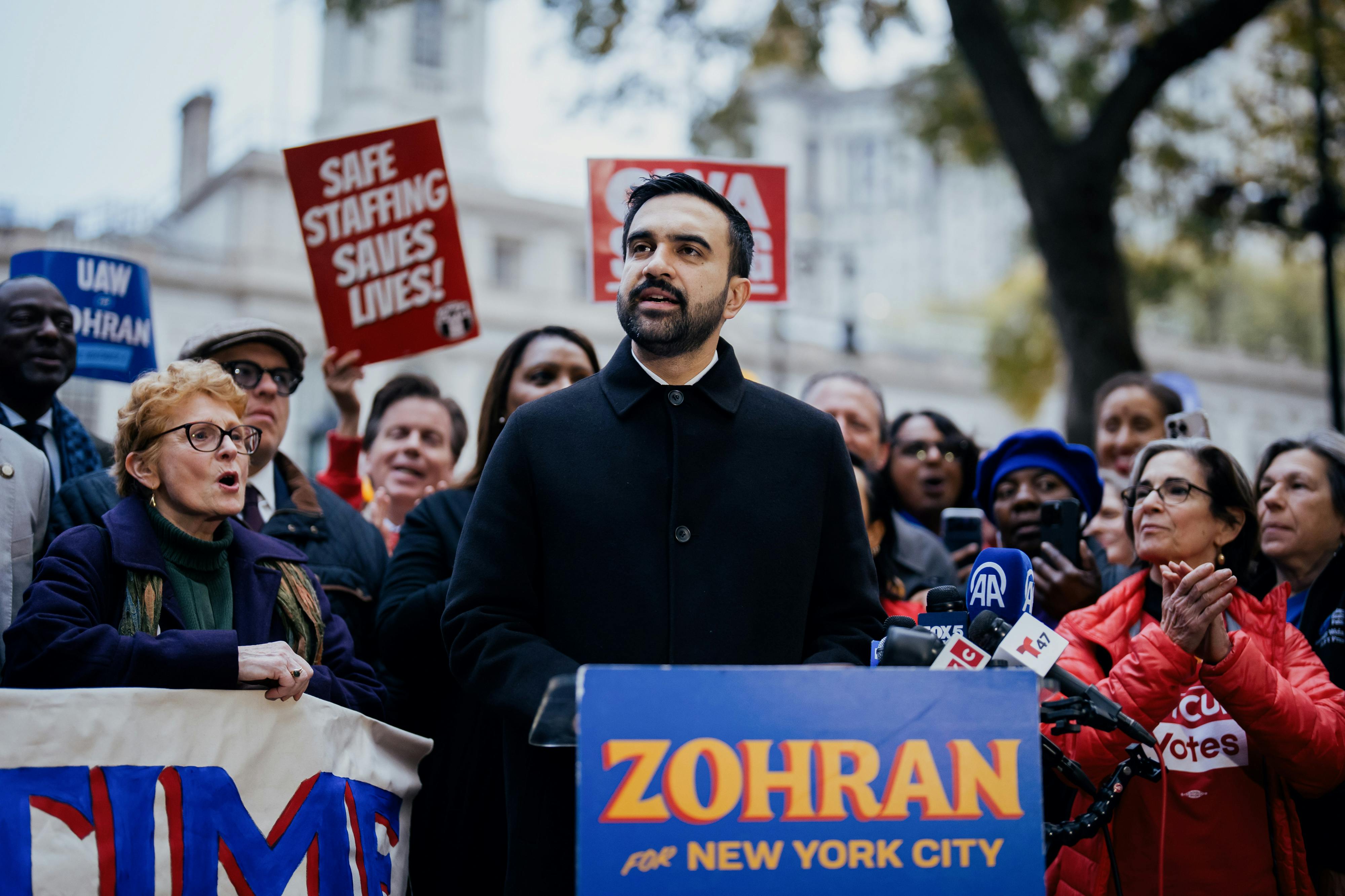 Zohran Mamdani speaks at a lectern as others behind him cheer, hold protest signs, and clap.