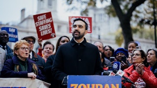 Zohran Mamdani speaks at a lectern as others behind him cheer, hold protest signs, and clap.