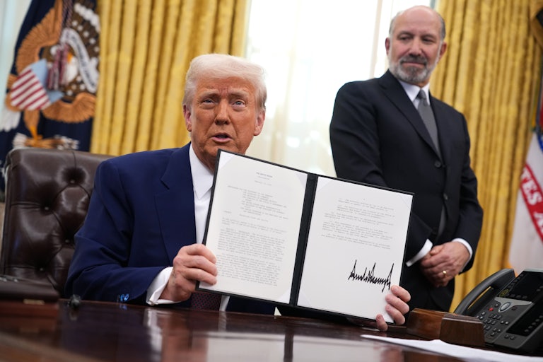 Commerce Secretary stands and watches as Donald Trump sits at his desk and holds up a signed executive order.