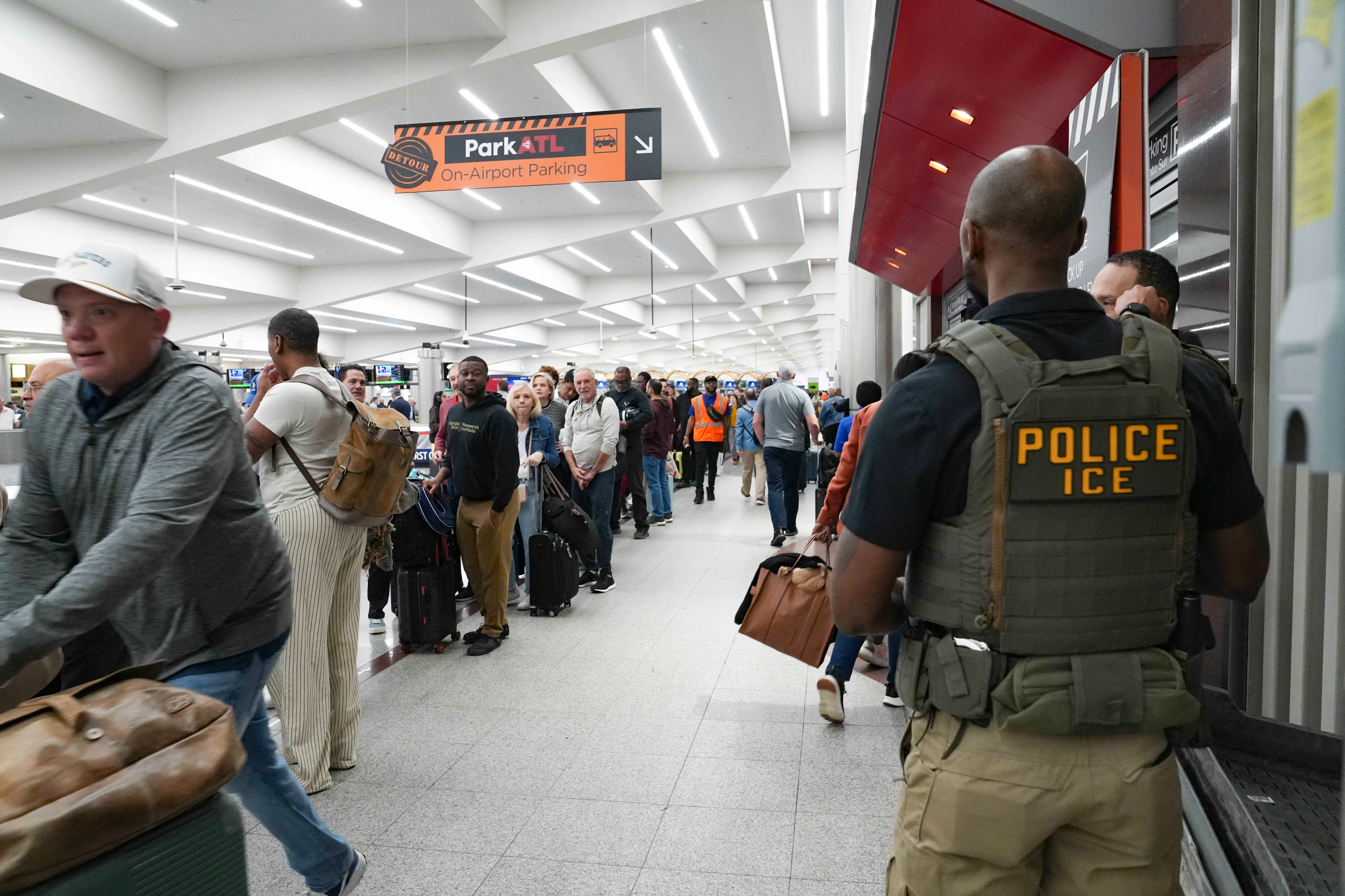 ICE agents stand looking at long lines in an airport terminal. One ICE agent is clearly wearing a flak jacket reading "Police ICE" is 
