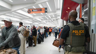 ICE agents stand looking at long lines in an airport terminal. One ICE agent is clearly wearing a flak jacket reading "Police ICE" is