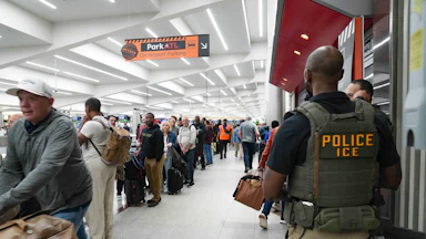 ICE agents stand looking at long lines in an airport terminal. One ICE agent is clearly wearing a flak jacket reading "Police ICE" is