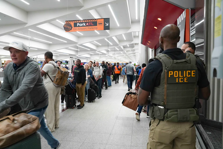 ICE agents stand looking at long lines in an airport terminal. One ICE agent is clearly wearing a flak jacket reading "Police ICE" is
