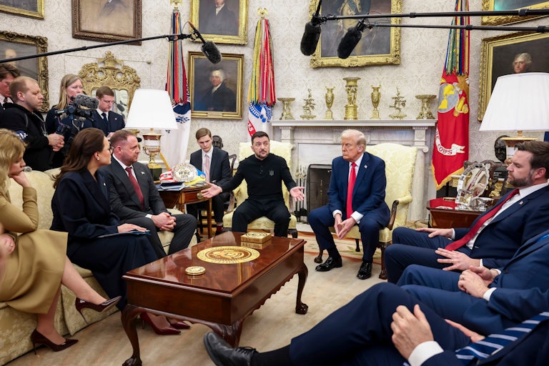 Reporters stand around the edge of the Oval Office. Volodymyr Zelenskiy, who holds his arms out the side, and Donald Trump sit in the middle