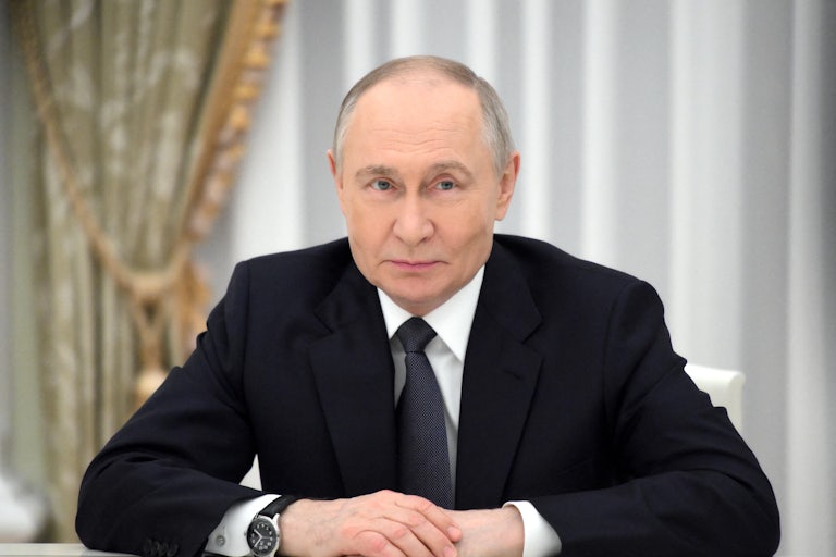 Russian President Vladimir Putin sits at a table with his hands folded on top of it
