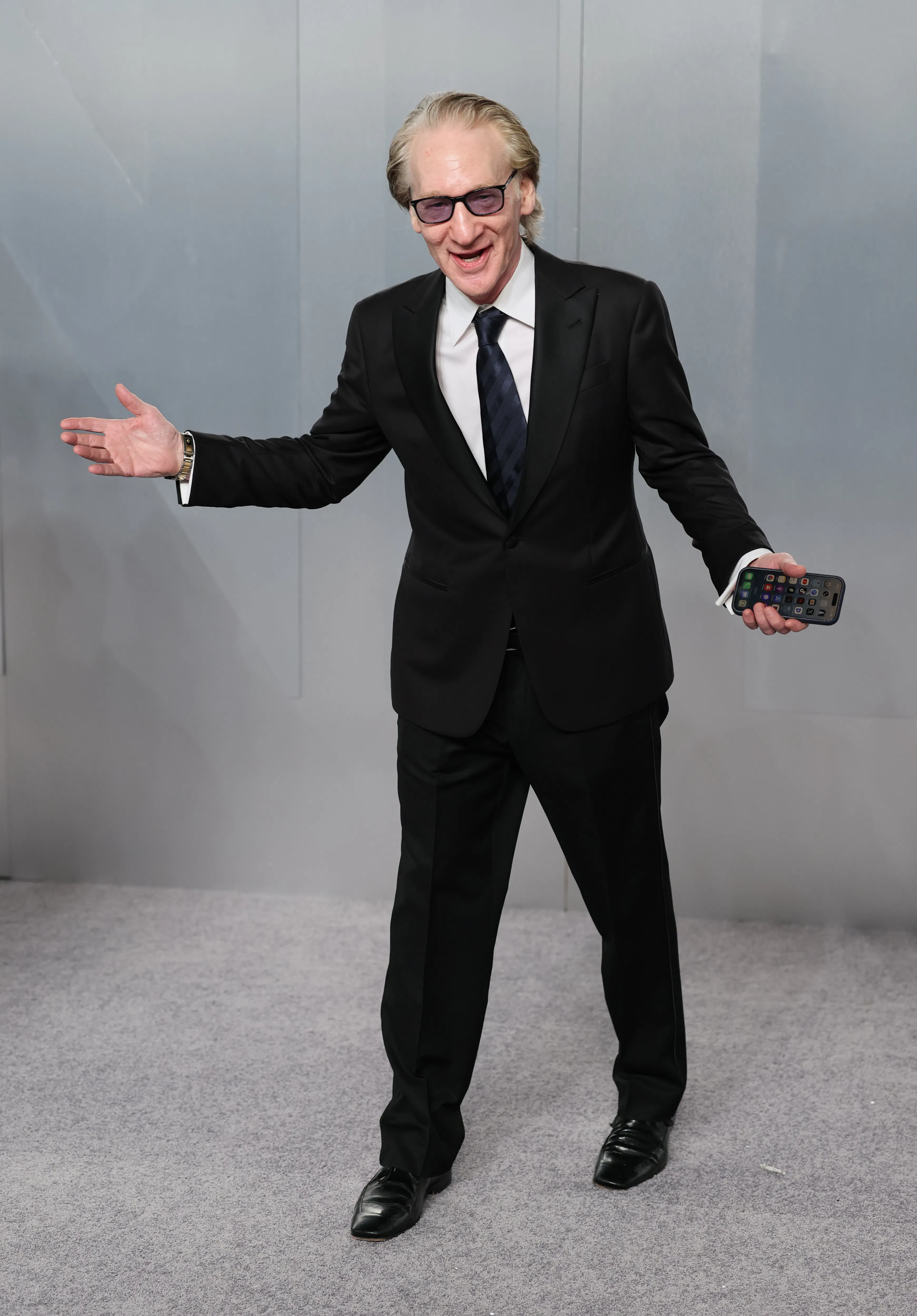 Bill Maher wears and tux and splays his arms out as if he's performing, while holding a phone in one hand