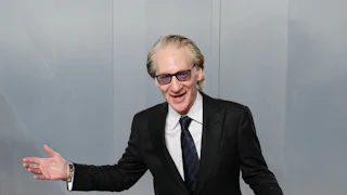 Bill Maher wears and tux and splays his arms out as if he's performing, while holding a phone in one hand