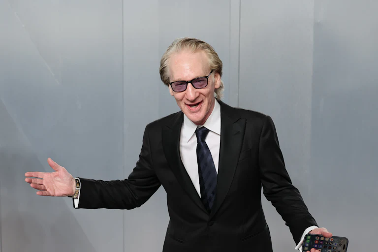 Bill Maher wears and tux and splays his arms out as if he's performing, while holding a phone in one hand