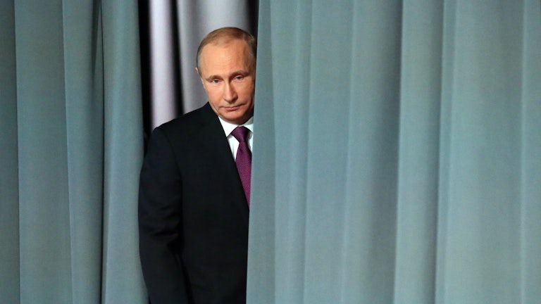 Russian President Vladimir Putin peeks out from behind a grey curtain.