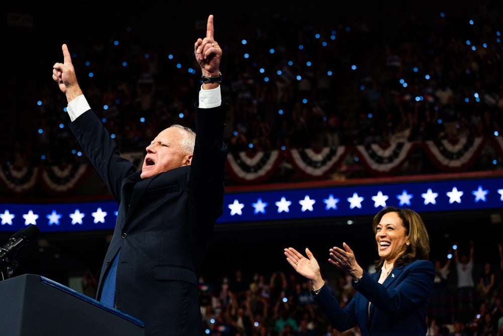 Tim Walz points at the sky with both hands as Kamala Harris smiles and applauds.