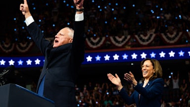 Tim Walz points at the sky with both hands as Kamala Harris smiles and applauds.
