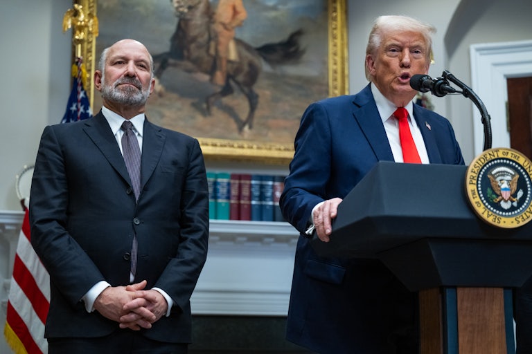 Howard Lutnick stands next to Donald Trump, who speaks at a podium in the Oval Office