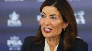 Kathy Hochul speaks in front of a blue background with a pattern.