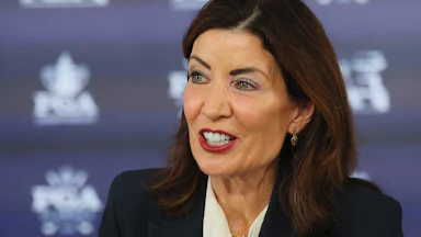 Kathy Hochul speaks in front of a blue background with a pattern.