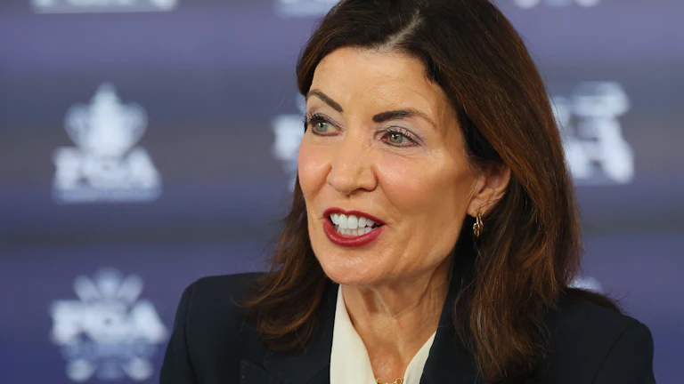 Kathy Hochul speaks in front of a blue background with a pattern.