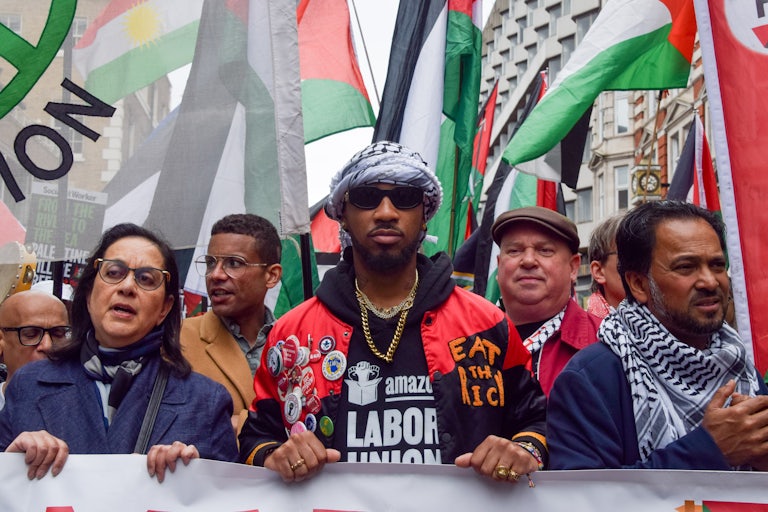 Chris Smalls wears a shirt that says Amazon Labor Union and a jacket that says Eat the Rich as he holds a banner alongside other protesters. He wears a keffiyeh on his head. Lots of Palestinian flags are in the background.
