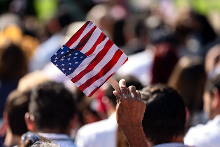 A brown hand holds up a paper U.S. flag fan amid a crowd of people in the sun.