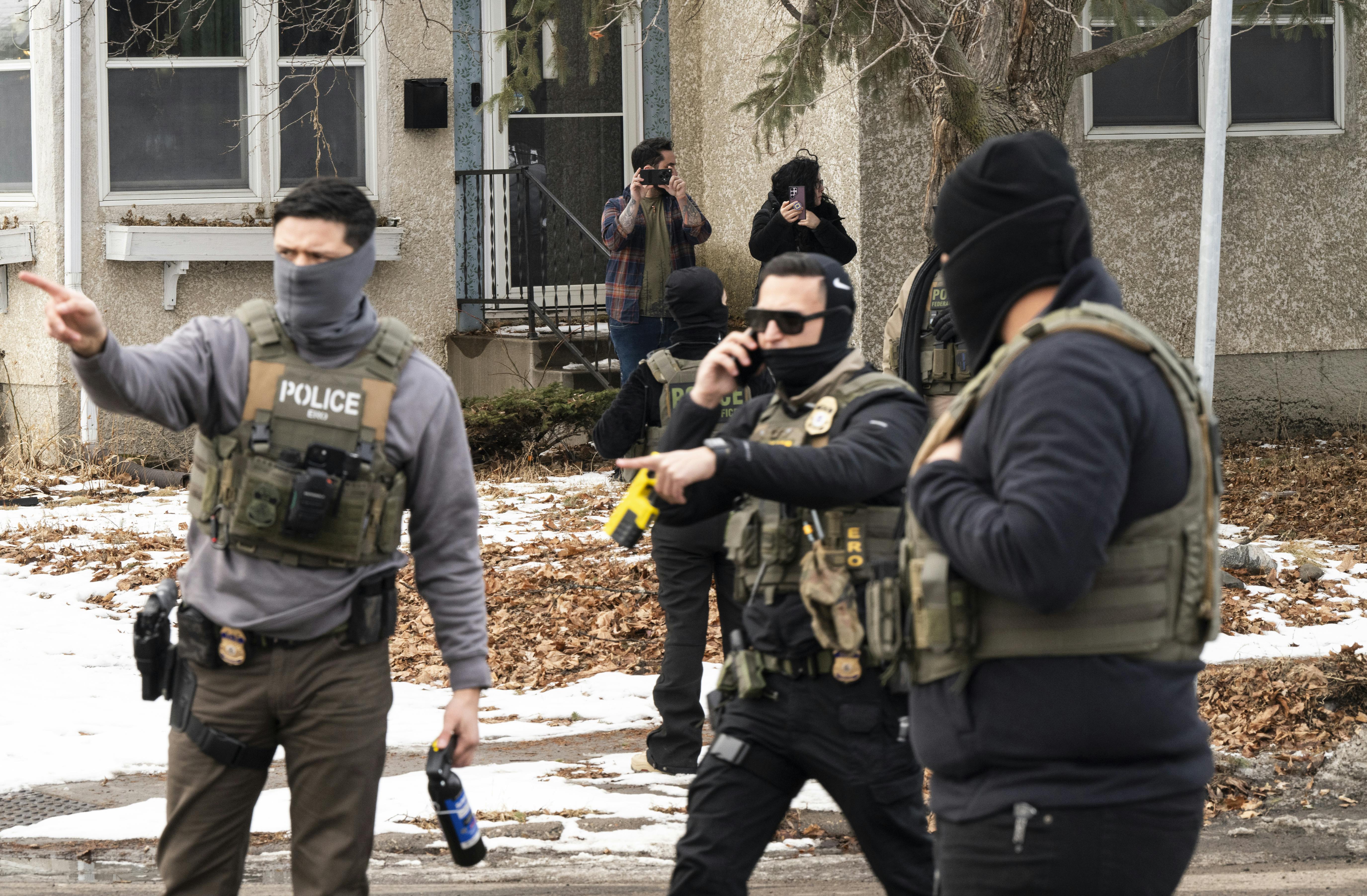 Federal immigration agents stand in a residential street. One of them points to the side.