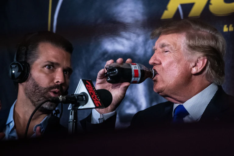 Donald Trump drinks a bottle of Diet Coke while sitting next to his son Donald Trump Jr.