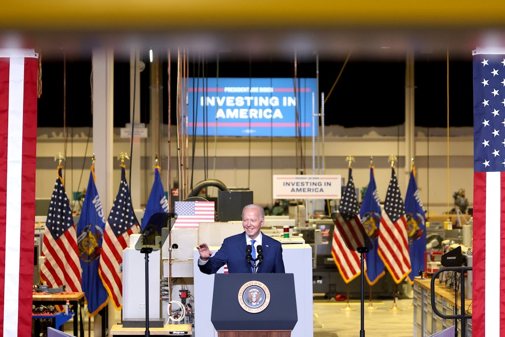 Joe Biden waves and smiles as he stands in front of several American flags and a large banner reading "INVESTING IN AMERICA."