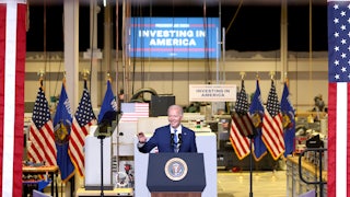 Joe Biden waves and smiles as he stands in front of several American flags and a large banner reading "INVESTING IN AMERICA."