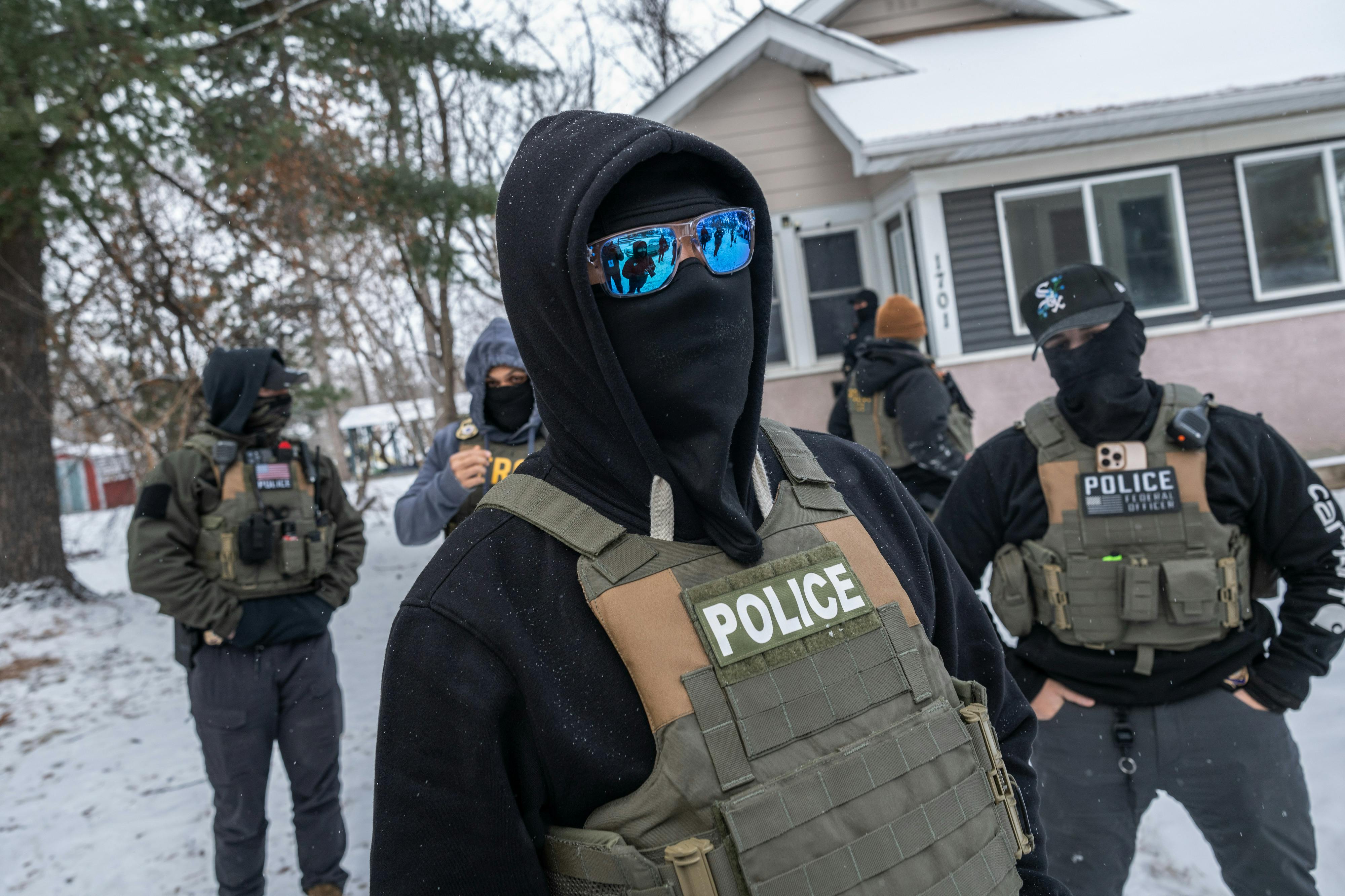 Masked federal immigration agents stand outside a house in the snow.