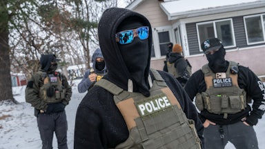 Masked federal immigration agents stand outside a house in the snow.