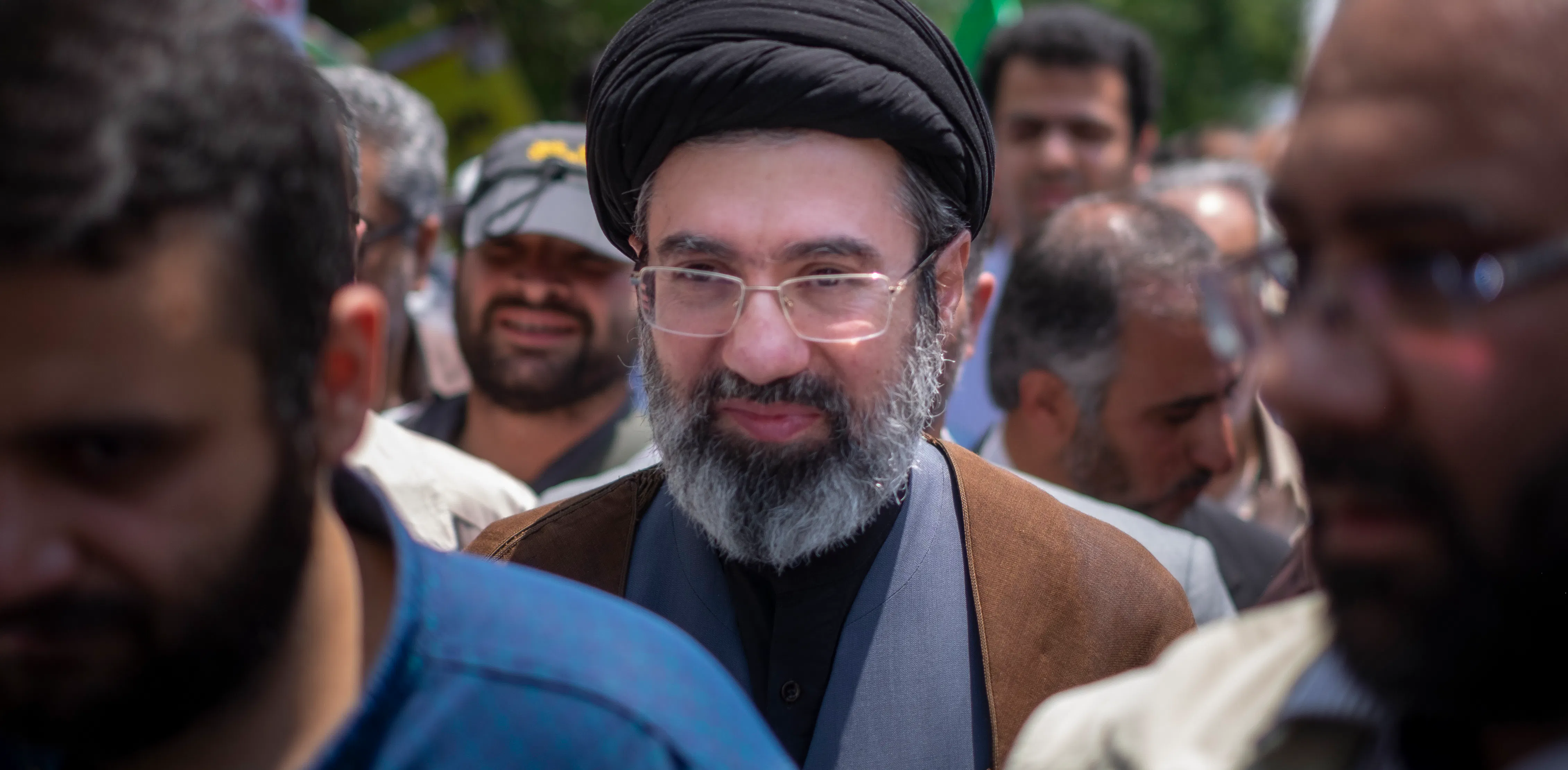 Ayatollah Mojtaba Khamenei, wearing a black turban and glasses, stands among a crowd of demonstrators. 