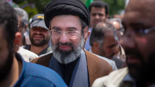 Ayatollah Mojtaba Khamenei, wearing a black turban and glasses, stands among a crowd of demonstrators.