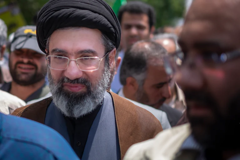 Ayatollah Mojtaba Khamenei, wearing a black turban and glasses, stands among a crowd of demonstrators.