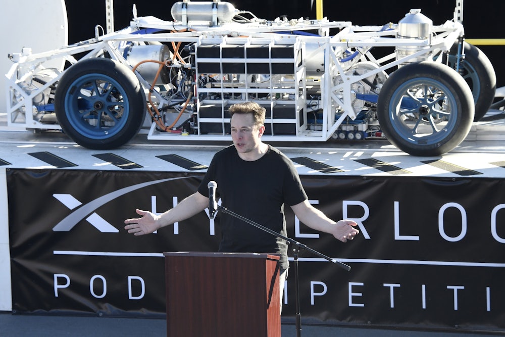 Elon Musk stands at a podium in front of a vehicle on a platform with a banner underneath reading HYPERLOOP POD COMPETITION.