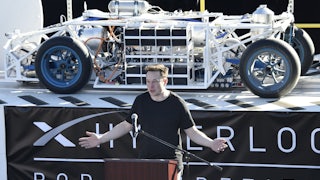 Elon Musk stands at a podium in front of a vehicle on a platform with a banner underneath reading HYPERLOOP POD COMPETITION.