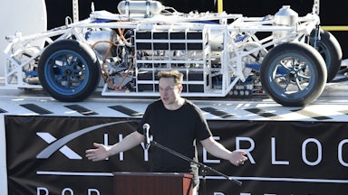 Elon Musk stands at a podium in front of a vehicle on a platform with a banner underneath reading HYPERLOOP POD COMPETITION.