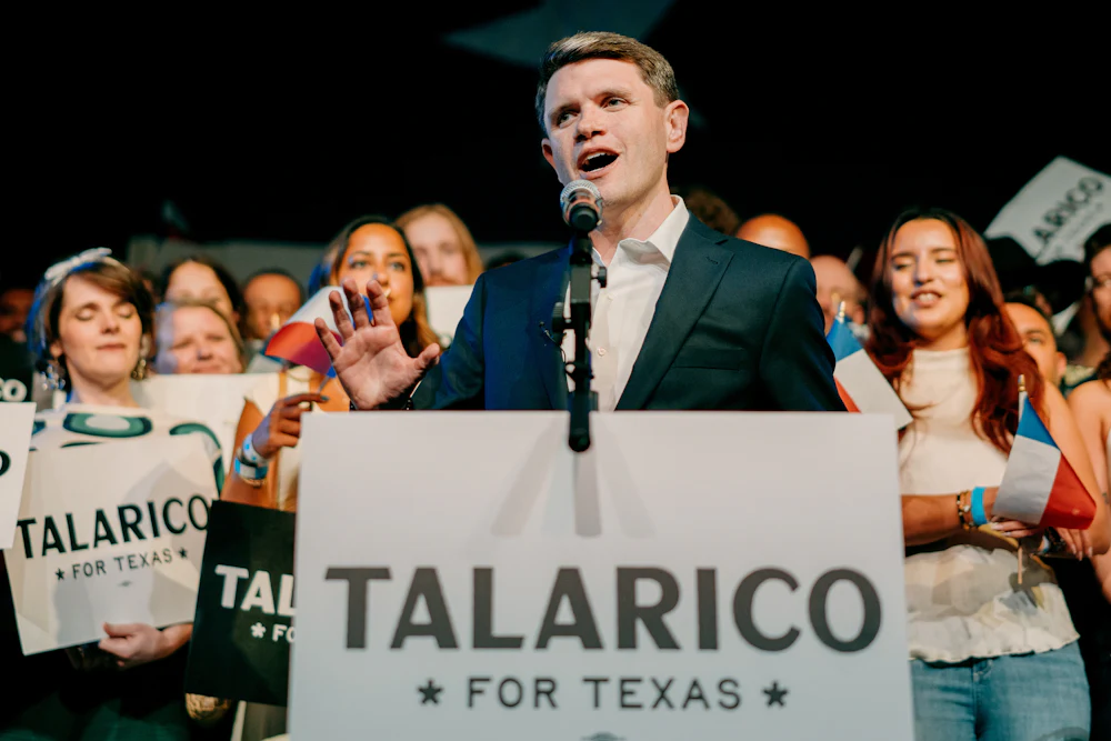 Talarico stands at a podium reading "TALARICO FOR TEXAS," speaking into a microphone as supporters stand behind him.