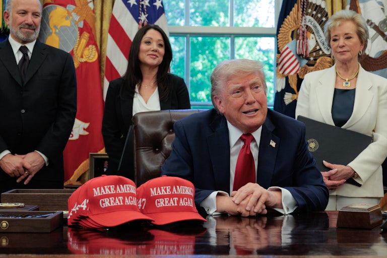 Donald Trump sits at his desk in the White House with two MAGA caps in front of him. Secretary of Commerce Howard Lutnick, Secretary of Labor Lori Chavez-DeRemer, and Secretary of Education Linda McMahon stand behind him.