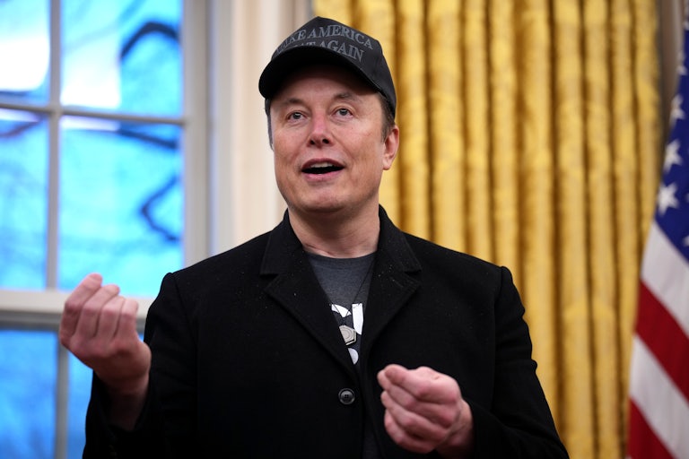 Elon Musk wears all black and a black MAGA cap while he speaks in the Oval Office