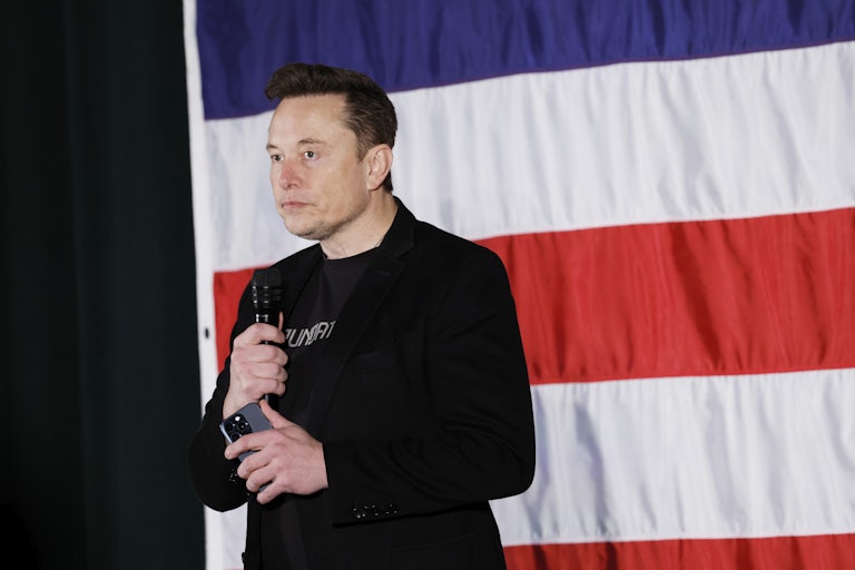 Elon Musk stands in front of a flag and grimaces