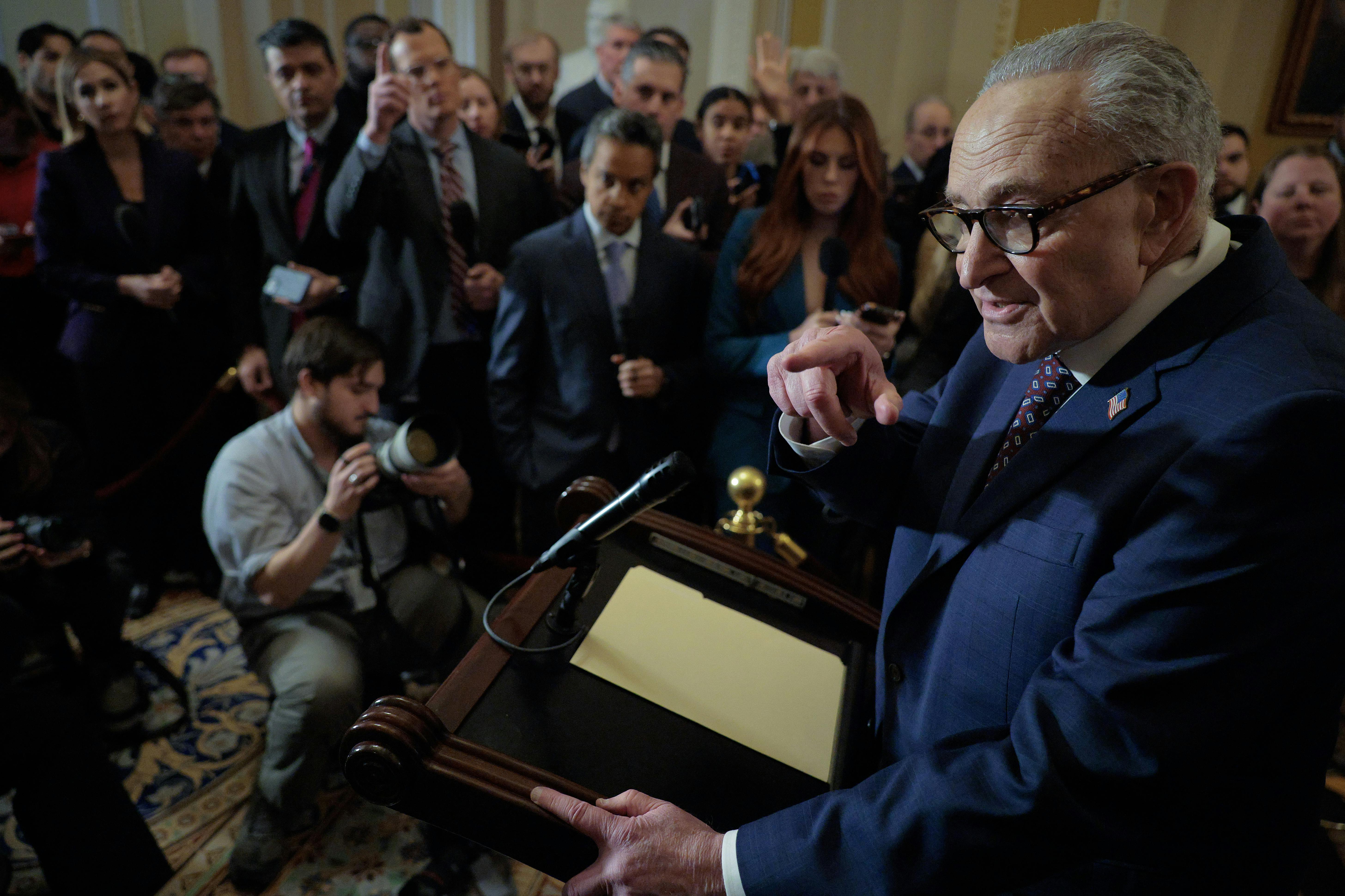 Chuck Schumer stands at a lectern in front of a group of reporters and points to one.