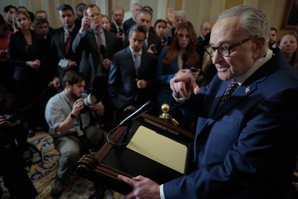 Chuck Schumer stands at a lectern in front of a group of reporters and points to one.