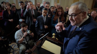 Chuck Schumer stands at a lectern in front of a group of reporters and points to one.