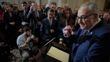 Chuck Schumer stands at a lectern in front of a group of reporters and points to one.