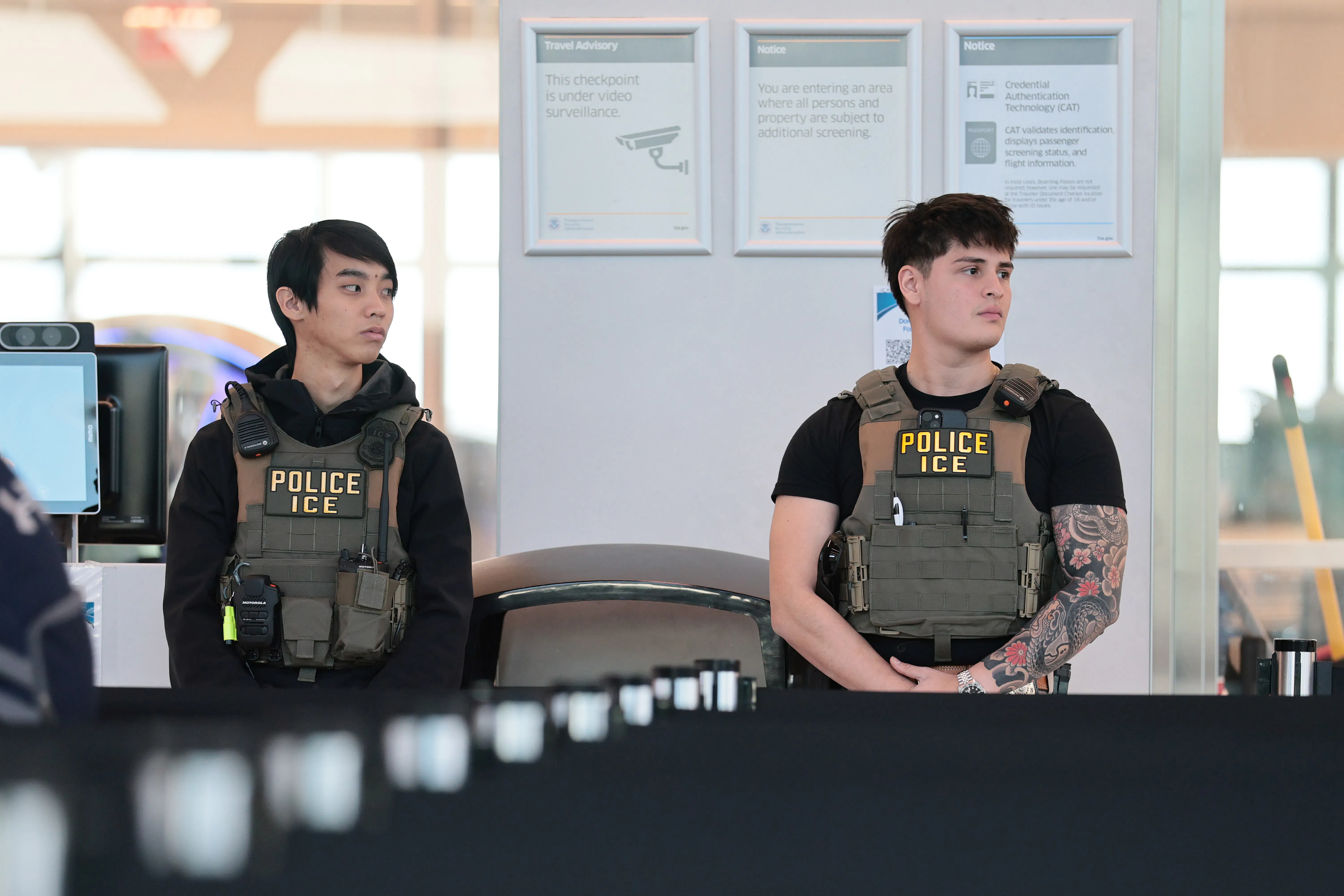 Two ICE agents wearing camoflage flak jackets, stand in an airport terminal.