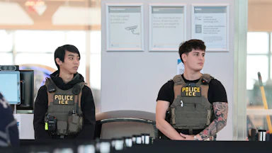 Two ICE agents wearing camoflage flak jackets, stand in an airport terminal.